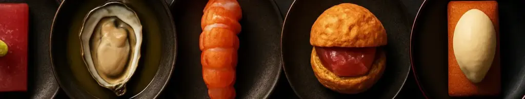 Seks japansk-franske anretninger på mørke tallerkener side om side, set lige ovenfra.