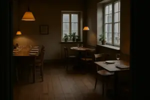 Et dansk restaurantlokale set fra døren med flere forskellige borde i samme rum: et langbord dækket til en stor familie, et lille tomandsbord ved vinduet, en enkelt kuvert med en bog ved siden af og et hjørnebord med en hundeskål på gulvet.