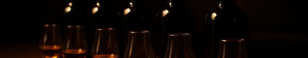 Mørke flasker med whisky og rom samt smagsglas med ravfarvet spiritus på en mørk bardisk