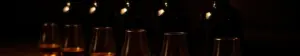 Mørke flasker med whisky og rom samt smagsglas med ravfarvet spiritus på en mørk bardisk